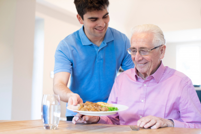 Male Care Assistant Serves Meal To Senior Male Seated At Table