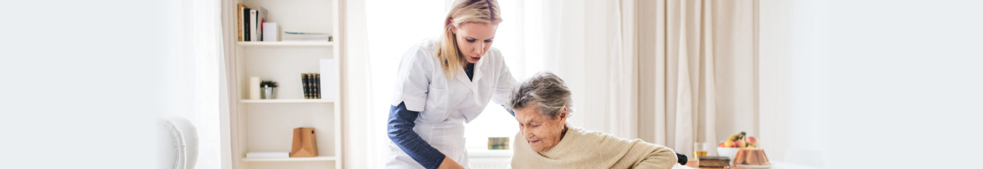 A young health visitor helping a senior women to stand up from a wheelchair at home.